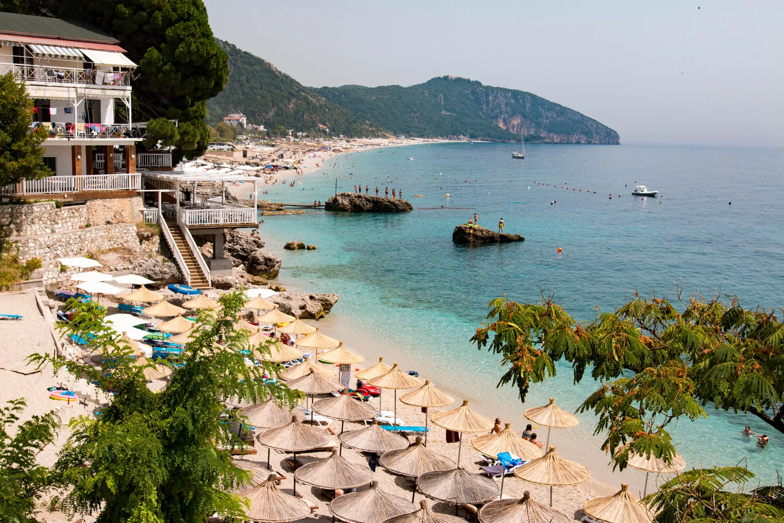 Dhërmi beach on the Albanian Riviera with thatched umbrellas, clear turquoise water and a green headland in the distance
