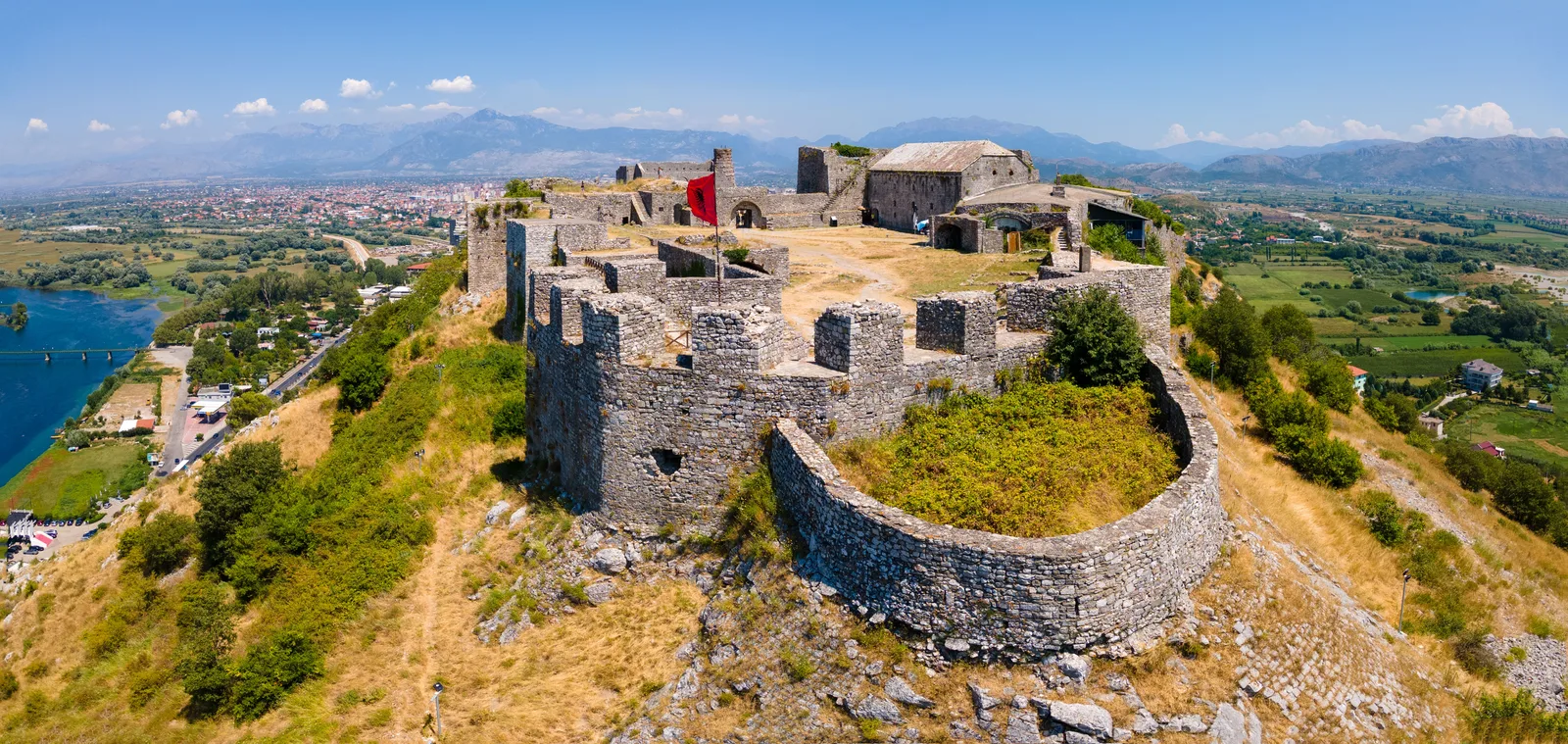 Aerial panorama of Rozafa Castle above Shkodër, Albania, with the Albanian flag flying over stone ramparts and rivers and mountains beyond