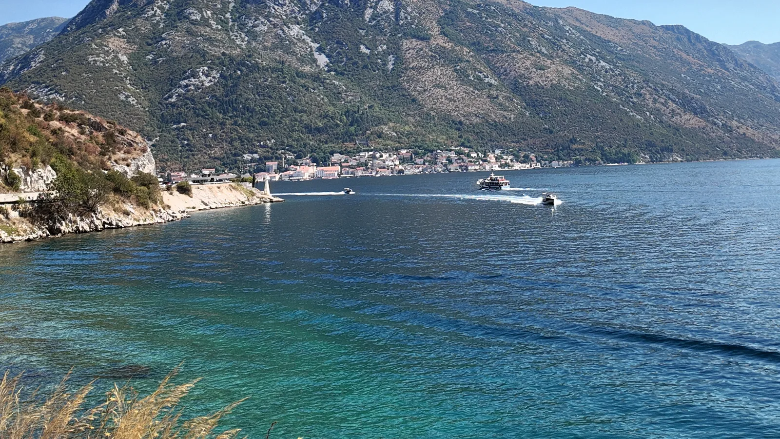 Perast, gesehen über die Meerenge von Verige: Uferfront und schlanker Glockenturm liegen am Fuß eines steilen Berghangs, ein Ausflugsboot und ein Schnellboot queren den Kanal, und ein kleines weißes Leuchtfeuer markiert die vordere Landspitze