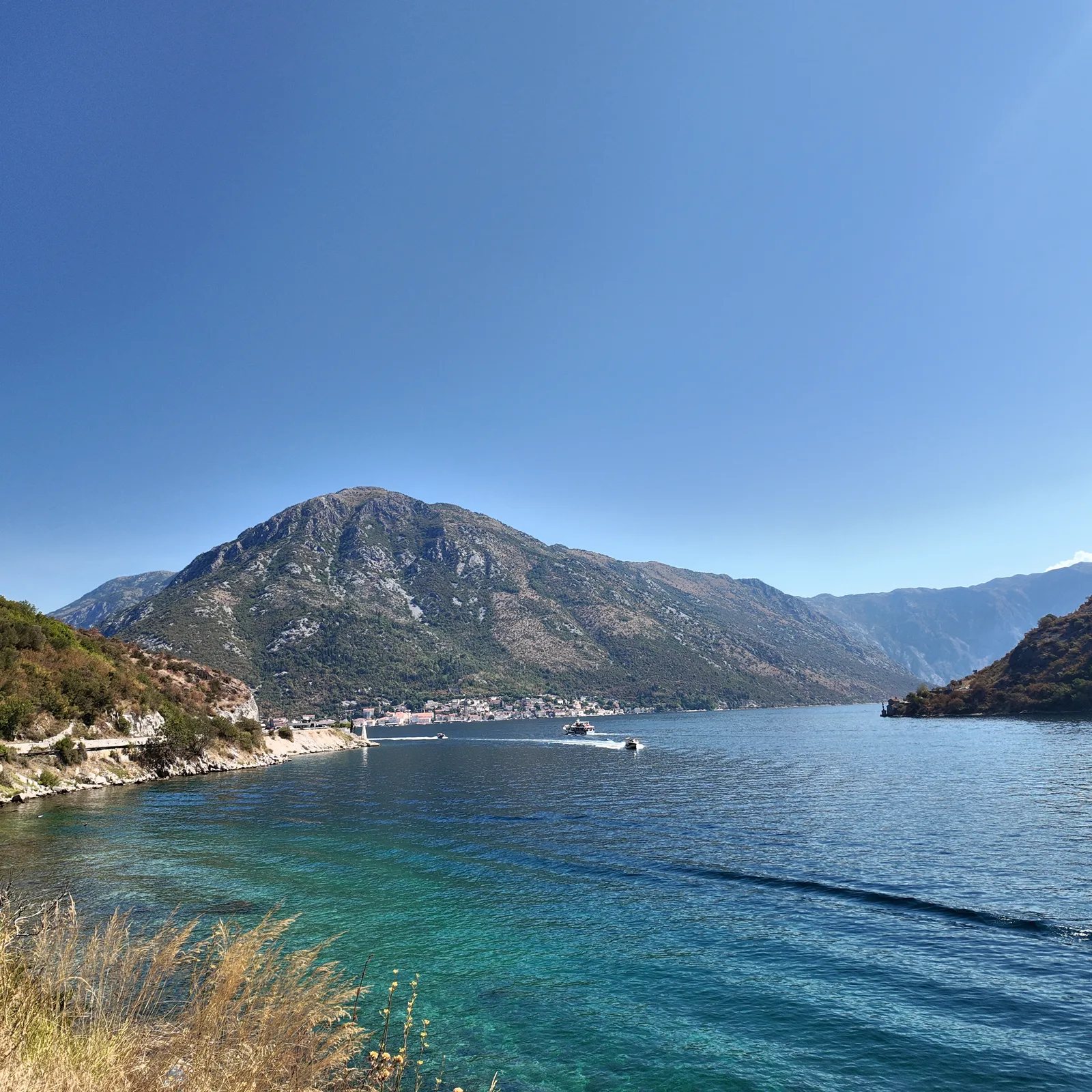 Die Meerenge von Verige, die schmalste Stelle der Bucht von Kotor, unter klarem Sommerhimmel: trocken-grüne Berghänge fallen zum blauen Wasser ab, kleine Boote queren den Kanal und ziehen weiße Kielwasserspuren, und jenseits der Bucht leuchtet die helle Uferfront von Perast