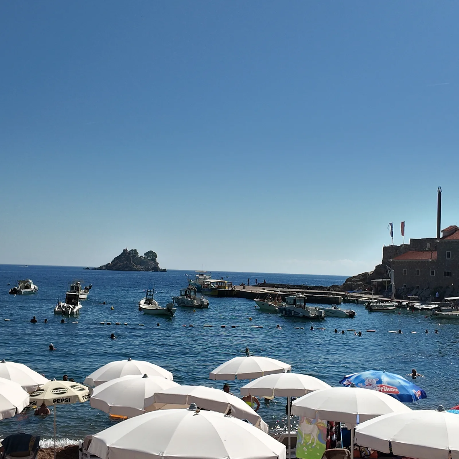 White beach umbrellas at the north end of Petrovac beach, swimmers inside the buoyed swimming lanes, small boats moored off the stone pier, the Castello fortress on its rock at right and the offshore islets on the horizon at left
