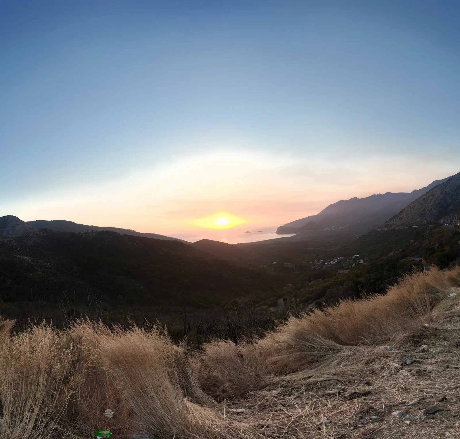Sunset over the Adriatic from the coast road above Buljarica, with the sun low over the sea, the long arc of Buljarica beach and its green plain below, and the Katič islets visible beyond the headland toward Petrovac