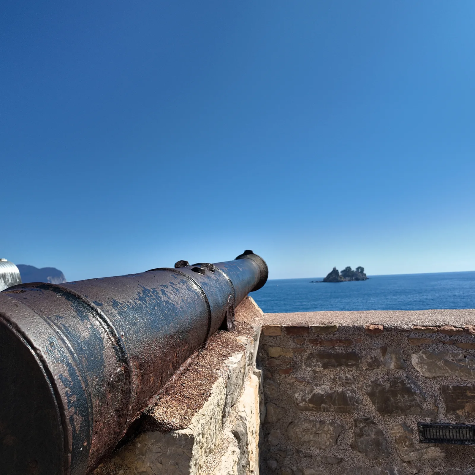 A rusted iron cannon on the stone parapet of the Castello fortress in Petrovac, pointing out over a deep blue sea with the wooded offshore islets in the distance at right