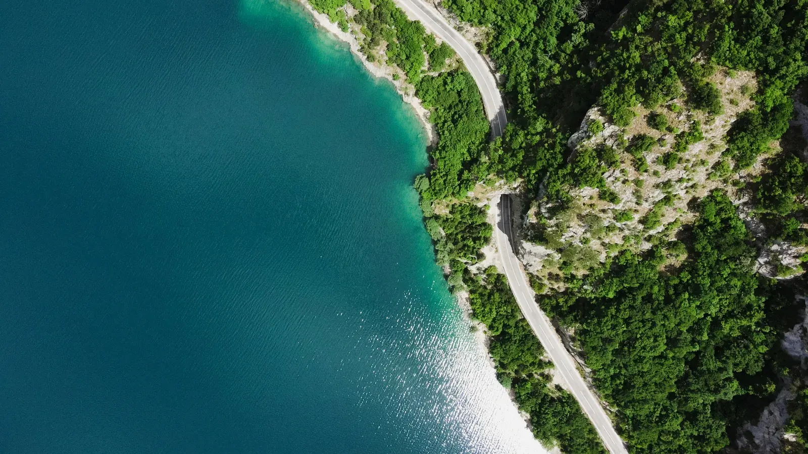 Aerial view of a coastal road cut into forested cliffs above turquoise water, passing through a short tunnel in the rock