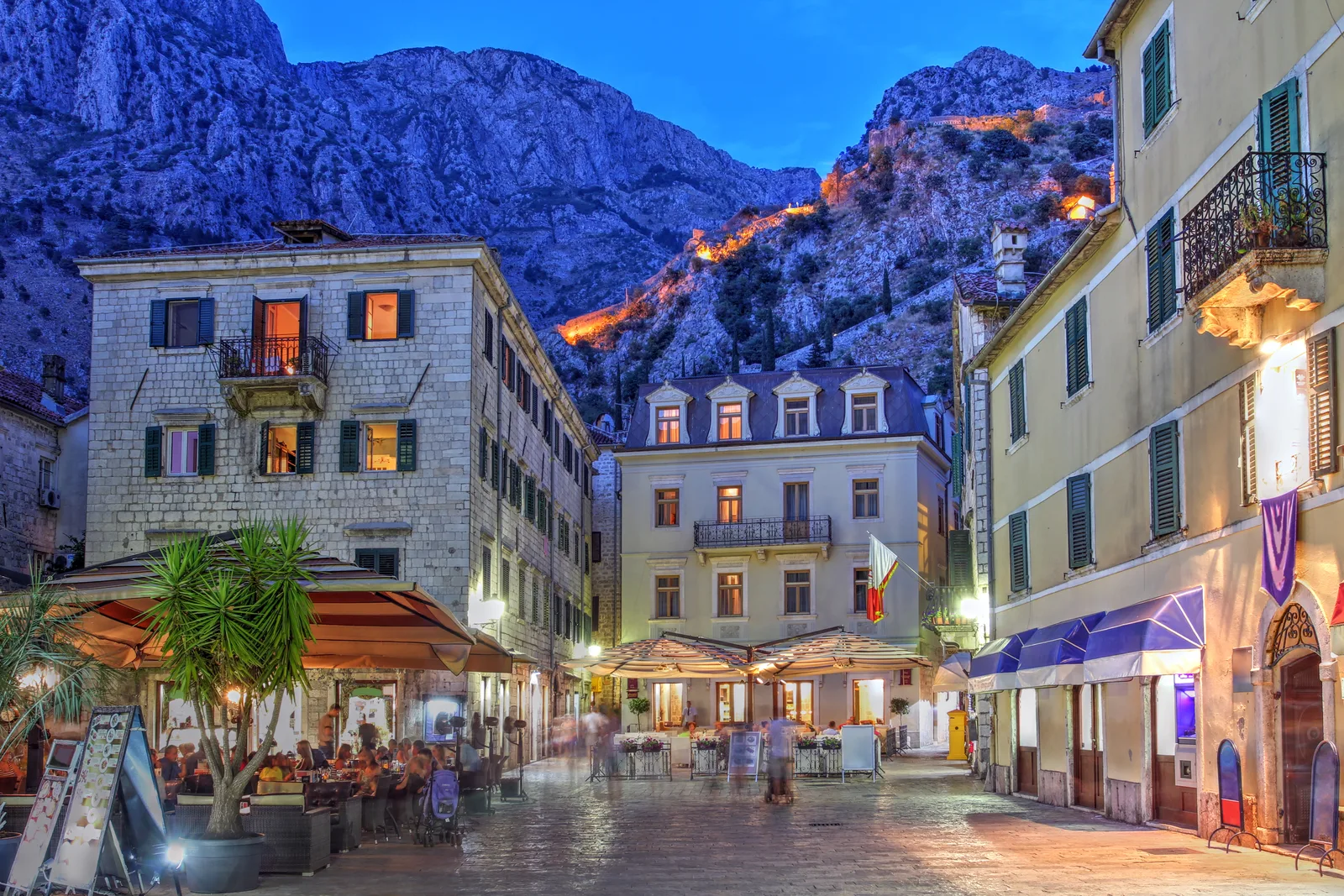 Plaza de Kotor Old Town por la noche con restaurantes iluminados y murallas de la ciudad iluminadas subiendo la montaña