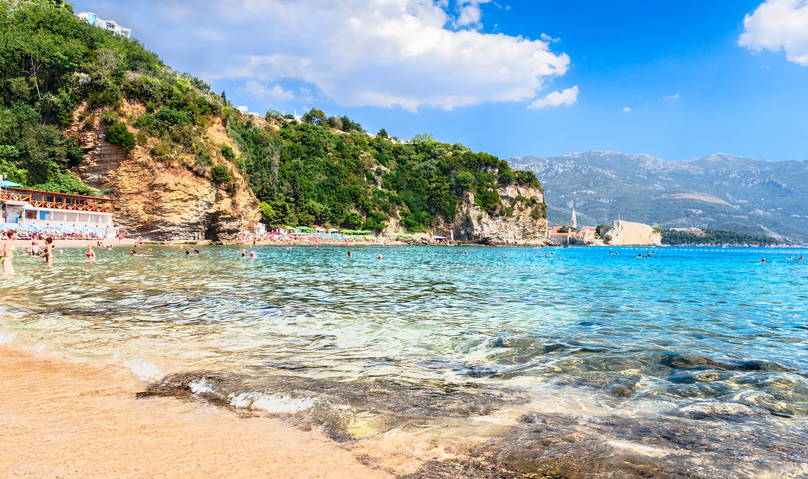 Mogren Beach cerca de Budva — arena dorada y un bar de playa bajo los acantilados, con Budva Old Town a la distancia
