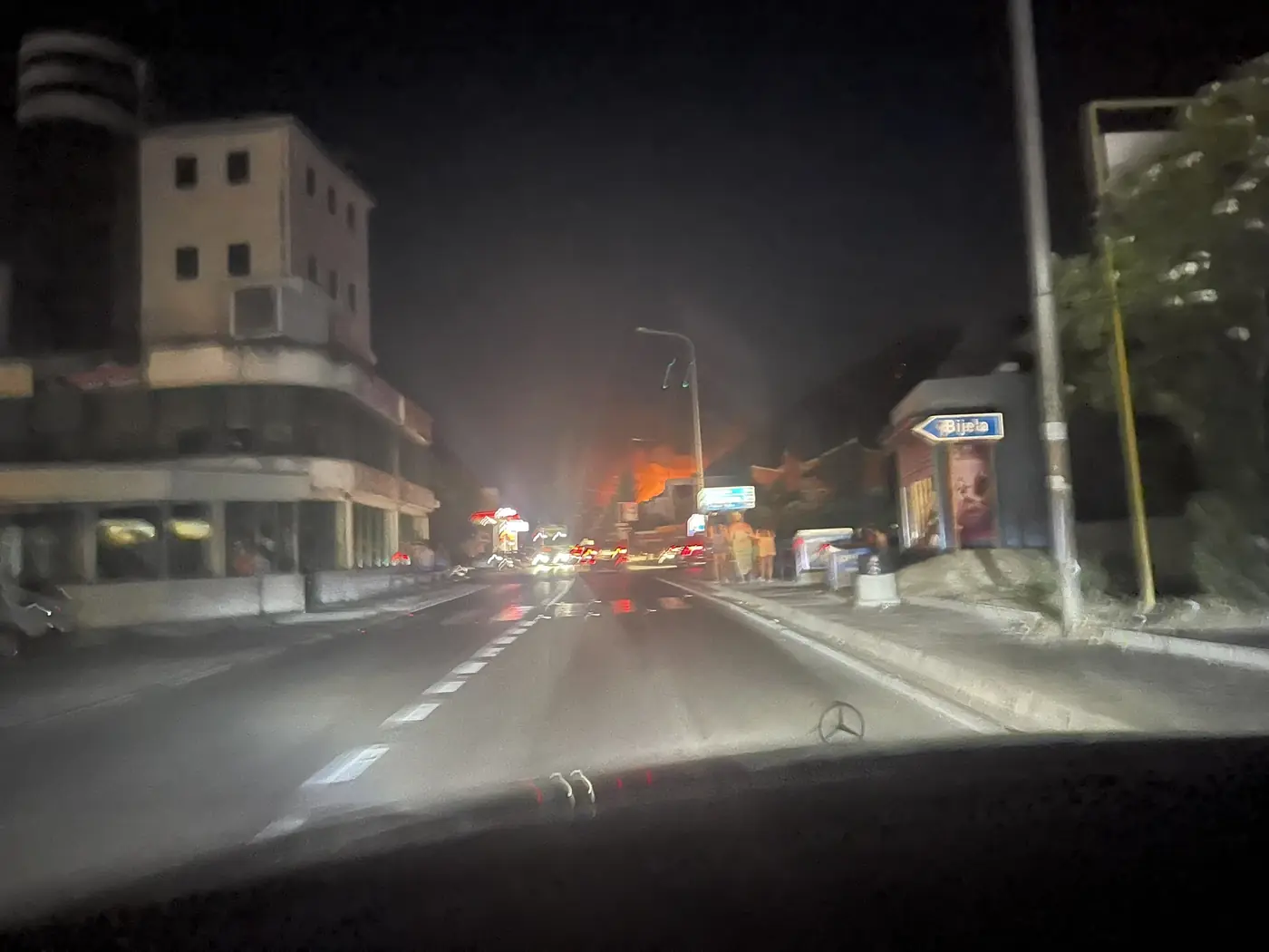 Traffic moving normally on the Adriatic highway at Bijela at night, past a road sign for Bijela, with the glow of the hillside fire ahead