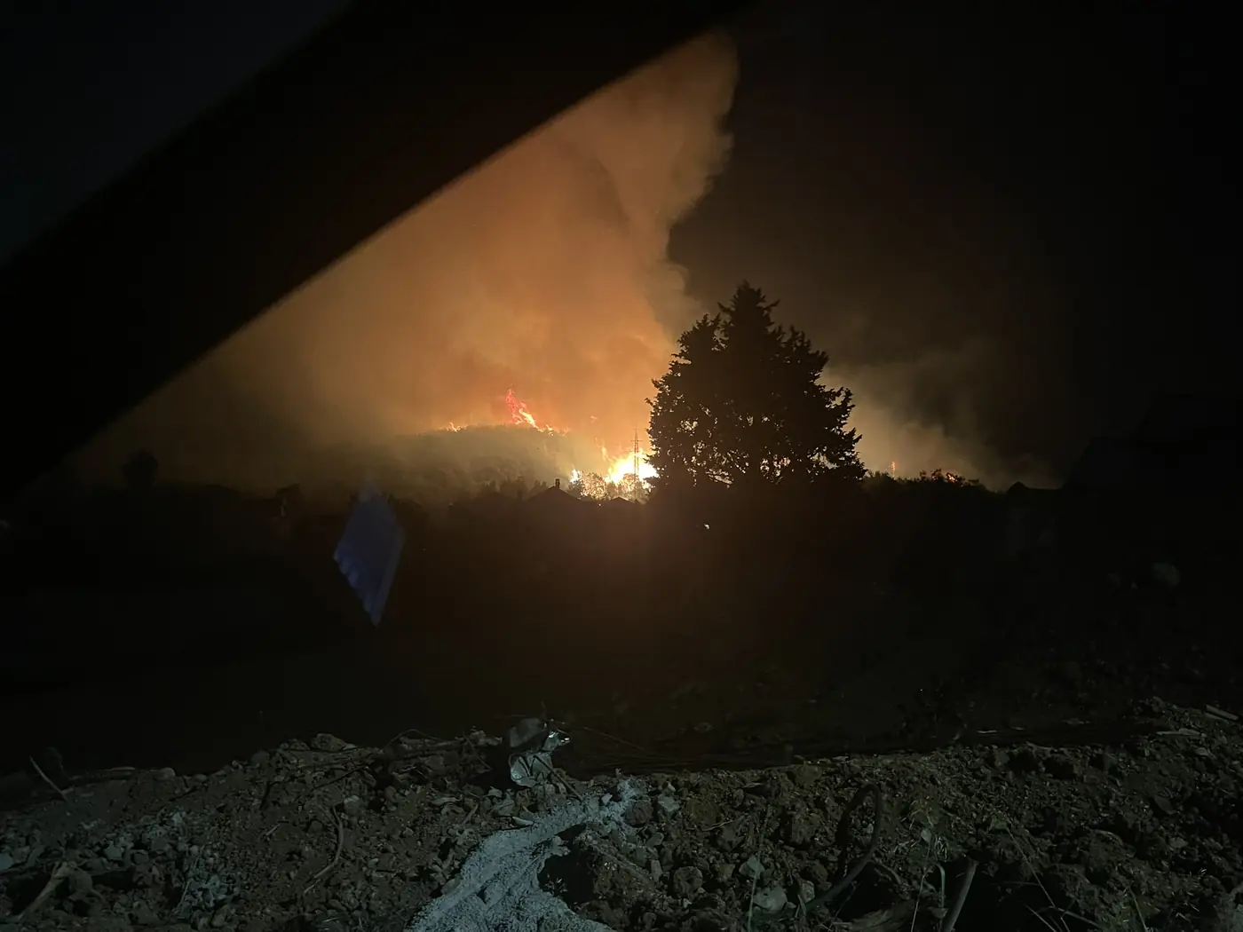 Fire burning above the rooftops of Đenovići at night, seen past the silhouette of a large tree