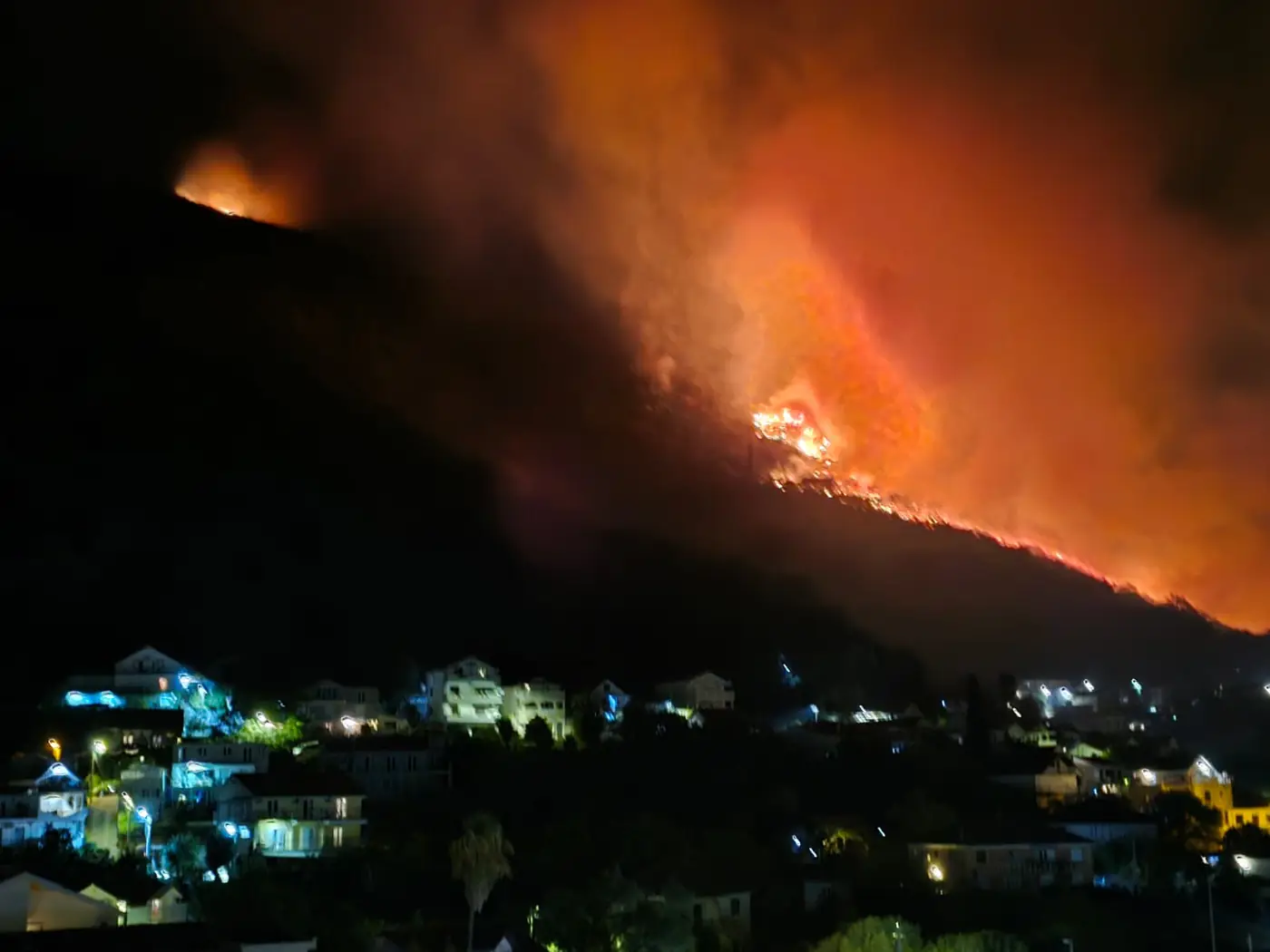 The Đenovići fire front burning along the ridge above the village at night, with the lit windows and street lamps of the houses below it untouched