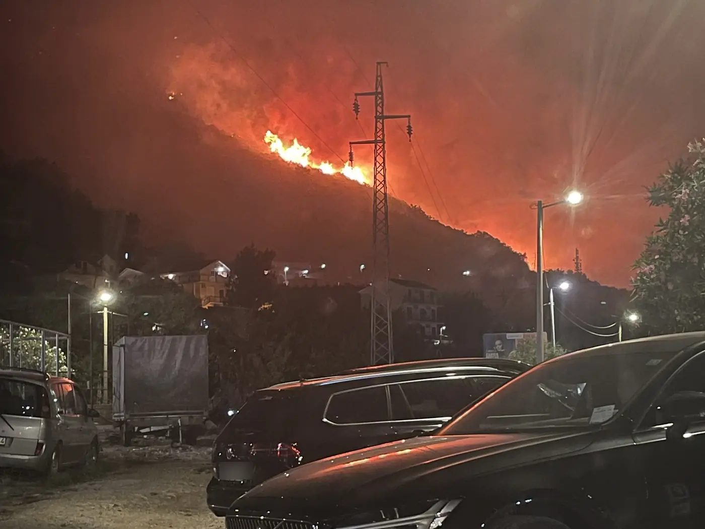 The burning ridge above Đenovići at night seen across a car park, a power pylon in silhouette, houses with their lights on below the fire and cars parked in the foreground