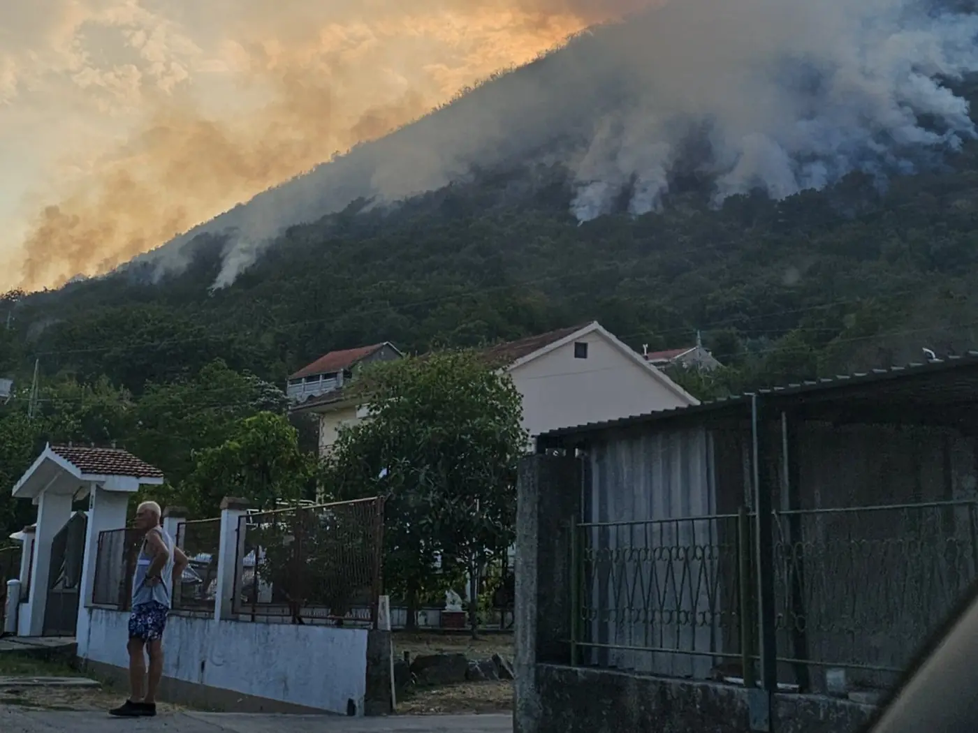 A resident stands at his garden gate in Đenovići with his hands on his hips, watching smoke and flame roll along the wooded hillside above the village roofs