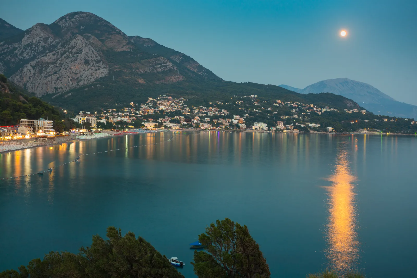 Sutomore a la hora azul, su costa iluminada reflejada en la tranquila bahía bajo la luna llena