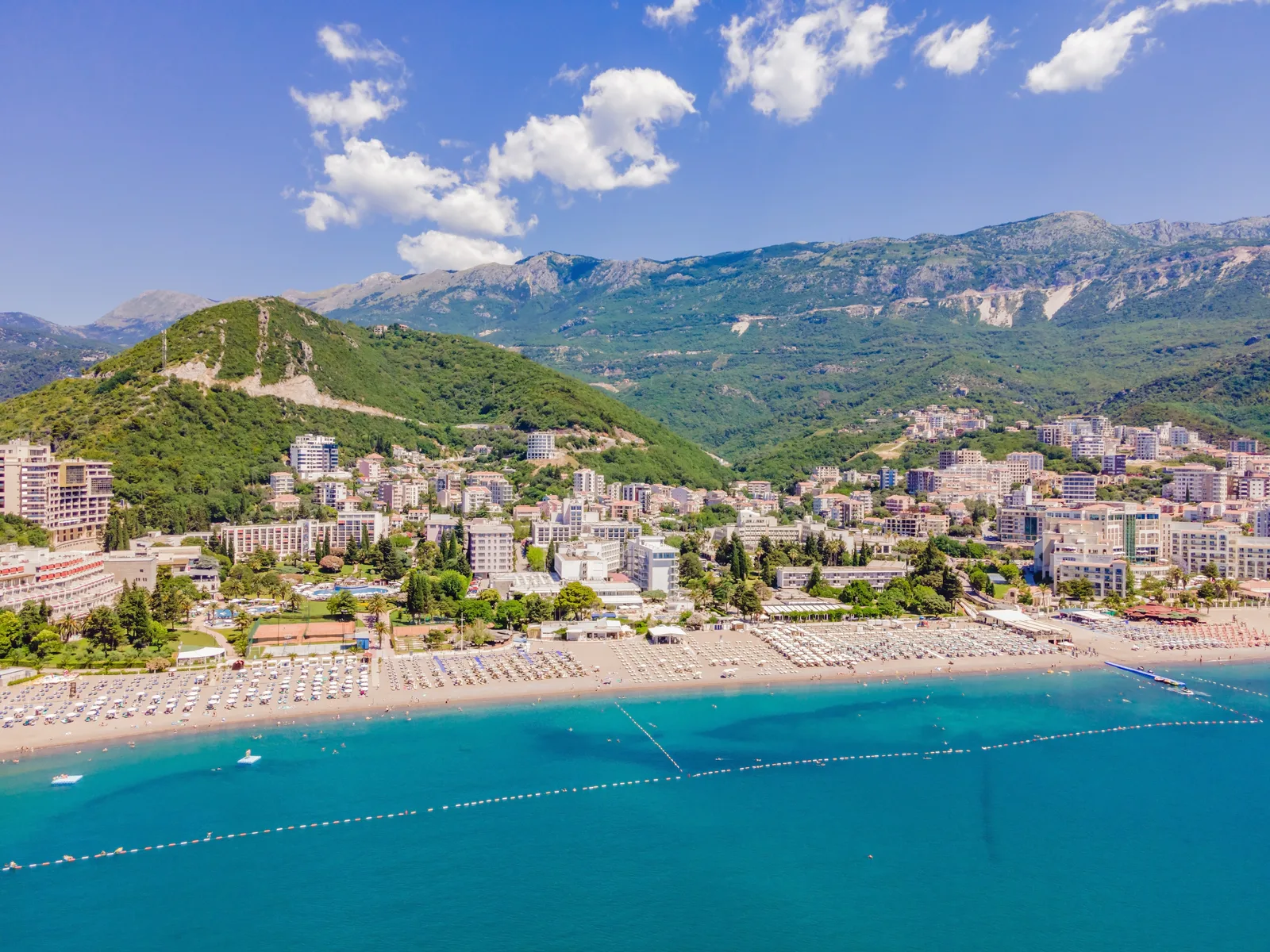 Vista aérea de los hoteles, jardines y playa llena de sombrillas de Becici por debajo de montañas verdes