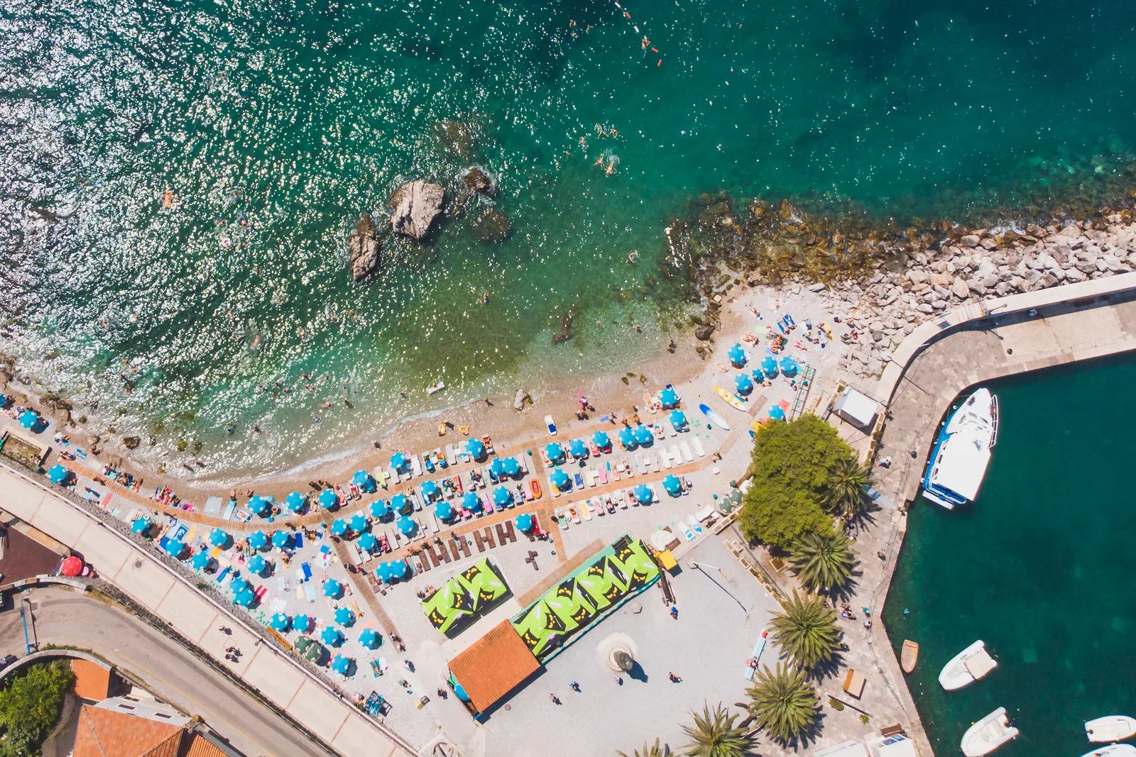 Aerial top-down of a Herceg Novi beach with blue umbrellas, a pier and moored boats