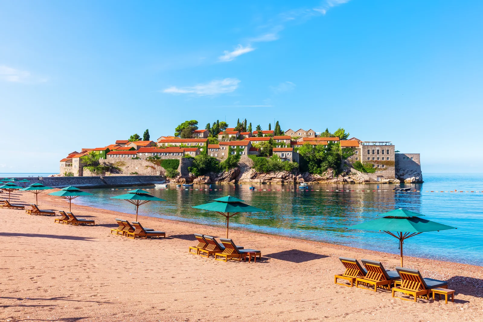 Green umbrellas and wooden loungers on the pink-sand beach facing Sveti Stefan island at morning