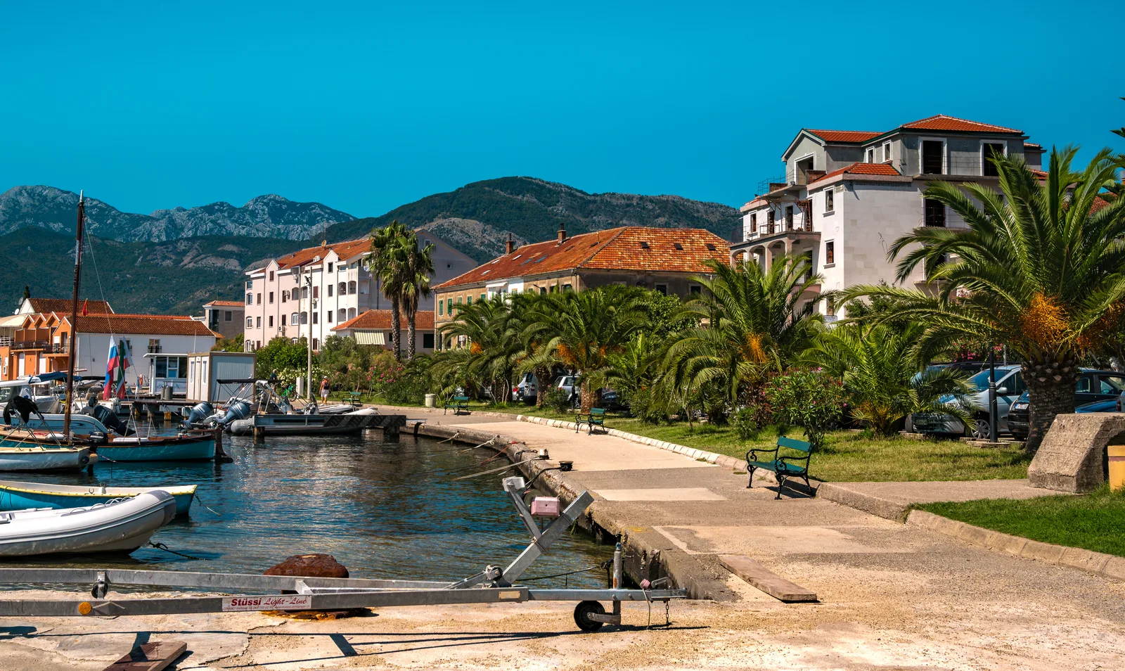 A quiet local waterfront with boats, palms and houses in Tivat, backed by mountains