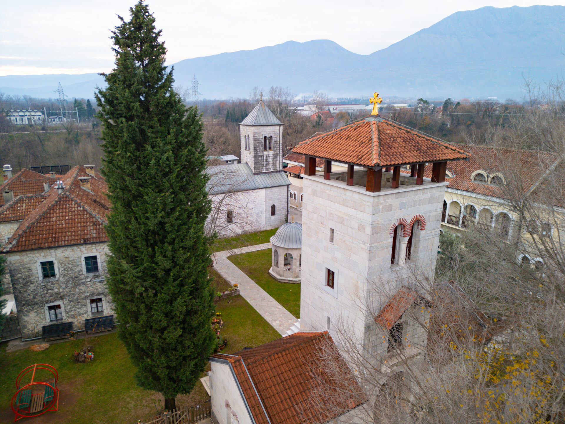 A historic Orthodox stone monastery with a tower and a tall cypress standing in the plain beneath rugged mountains near Danilovgrad