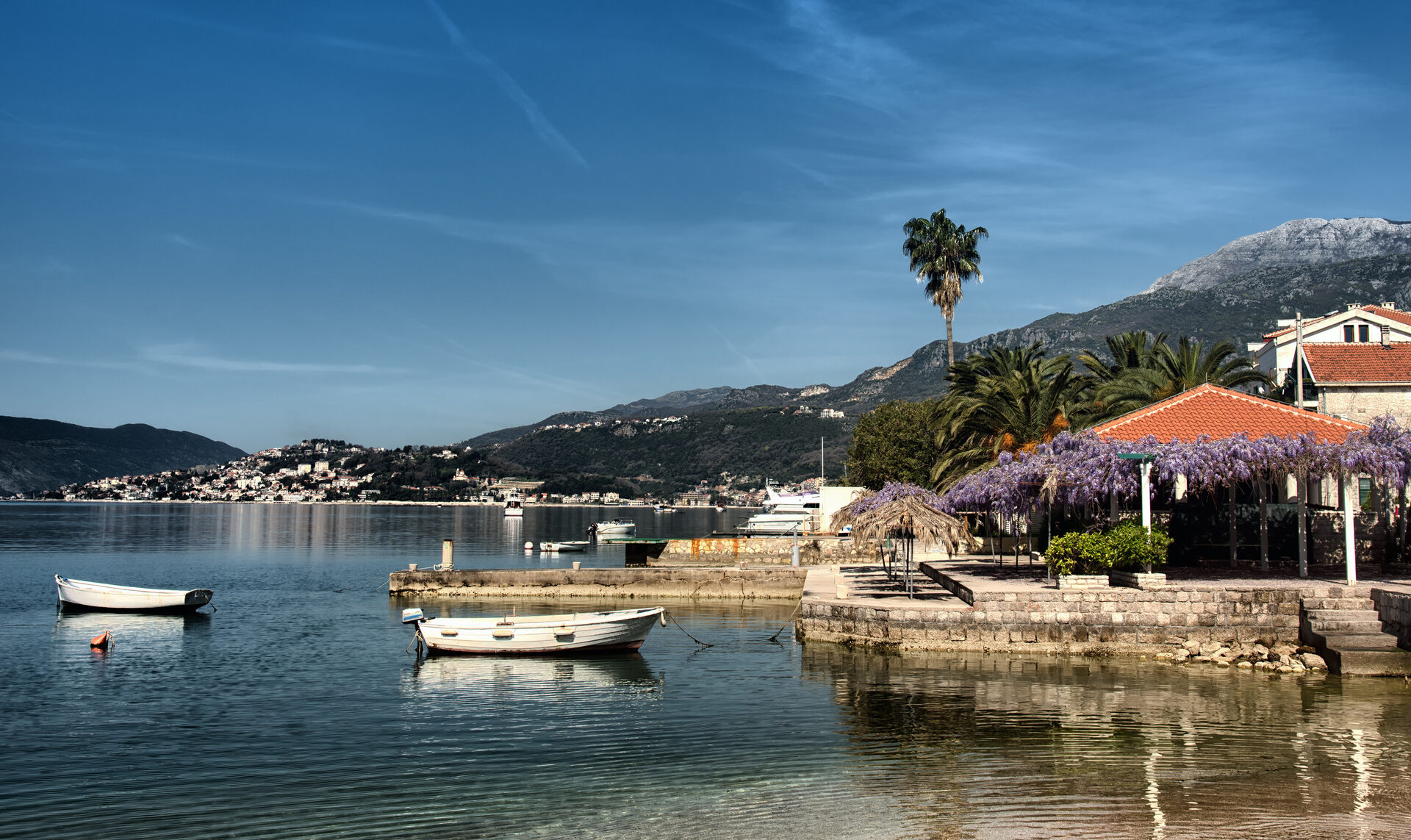A small boat moored in calm bay water beside a stone waterfront lined with palms